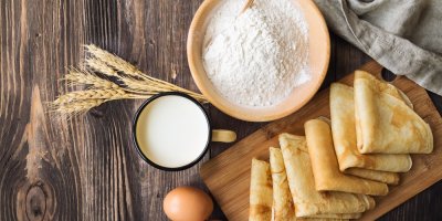 Několik palačinek, naskládaných na dřevěném prkénku, vedle miska s moukou a hrnek mléka