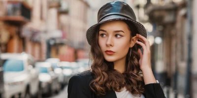 Mladá elegantní žena má na hlavě černý bucket hat z umělé kůže, pózuje v ulici velkoměsta