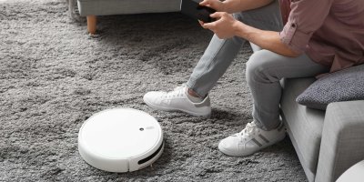 Robotický vysavač na koberci, vedle sedí člověk na sedačce