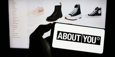 Člověk drží telefon s logem About You, v pozadí obrazovka s e-shopem