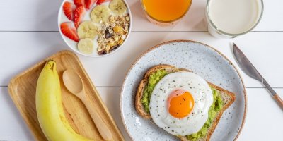 Toast s avokádem a vejcem, banán, miska jogurtu s granolou, banánem a jahodami,  sklenice džusu a mléka