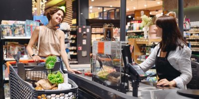 Mladá usmívající se zákaznice s nákupním vozíkem a paní pokladní u pokladny supermarketu
