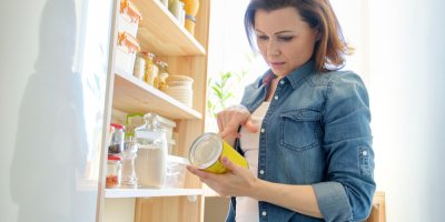 Žena stojí doma u regálu s potravinami a pročítá si informace na obalu konzervy, kterou drží v ruce