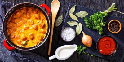 Vlevo párková omáčka nakkikastike v hrnci, vpravo vařečka, smetana, cibule, rajčatový protlak a koření