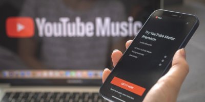 Žena drží telefon s aplikací YouTube Music, v pozadí obrazovka notebooku s logem YouTube Music