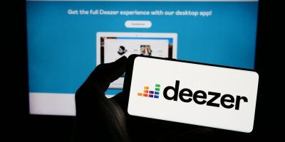 Člověk drží telefon s logem aplikace Deezer, v pozadí monitor s aplikací Deezer