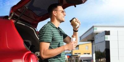 Muž pije kávu a v ruce drží párek v rohlíku a je opřený o červené auto