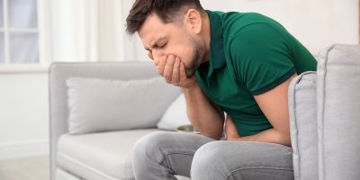 Muž sedí na sedačce a drží se za břicho, druhou ruku má před ústy