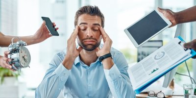 Muž v kanceláři se drží za hlavu, kolem něj ruce drží telefon, budík, tablet, složku