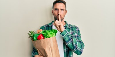 Muž drží papírovou tašku s nákupem a prstem u rtů žádá o ticho