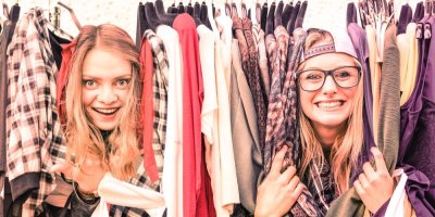 Dvě dívky žertovně vystrkují obličeje zpoza oblečení, které je vystavené na bleším trhu