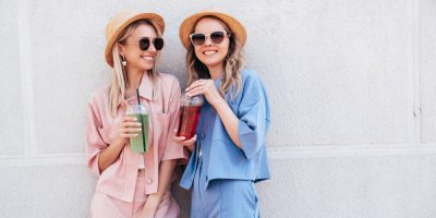 Dvě mladé usmívající se ženy v růžovém a modrém letním outfitu, s kloboukem a s brýlemi mají v ruce nápoj v kelímku