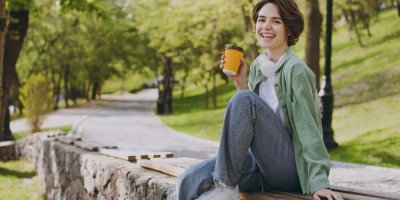 Mladá usmívající se žena sedí v parku a drží v ruce kelímek nápoje