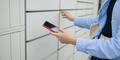 Žena drží v ruce telefon a stojí před boxem na balíčky