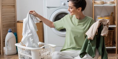 Žena má v koši na prádlo bílé oblečení, v ruce drží zelené oblečení