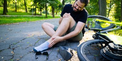 Muž s poraněným kotníkem sedí na cyklostezce v parku, vedle něj leží elektrokolo