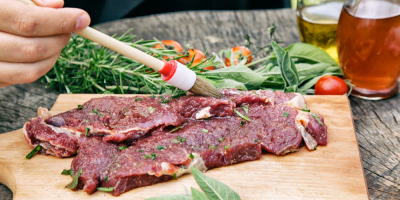 Záběr na ruku, která mašlovačkou potírá maso olejem s kořením