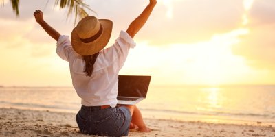 Mladá žena sedí s notebookem u oceánu a pozoruje západ slunce