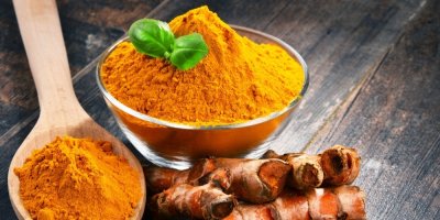 Skleněná miska s kurkumou, položená na stole. Vedle misky dřevěná lžíce s kurkumou a kurkumové oddenky