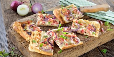 Alsaský cibulový koláč nakrájený na kousky, bylinky a červená cibule v pozadí