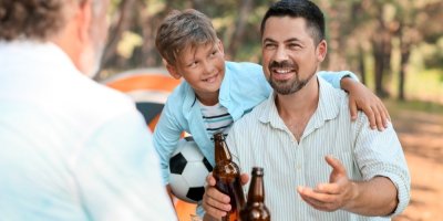 Muž drží lahev od piva, hovoří s dalším mužem, objímá ho kolem ramen jeho syn