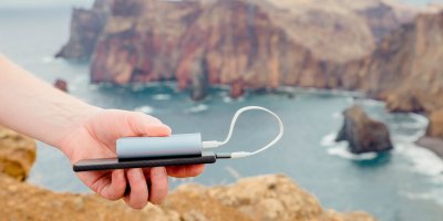 Ruka drží power banku nabíjející mobil, v pozadí je mořská zátoka