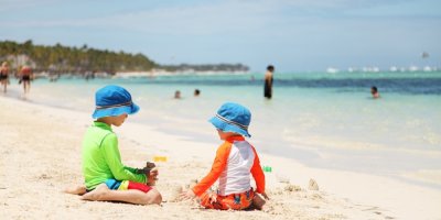 Dvě děti s kloboučky a v koupacích tričkách s dlouhými rukávy si hrají na pláži.