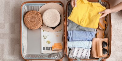 Otevřený kufr, do kterého člověk přidává košili. Jsou v něm kraťasy, klobouk, kabelka, brýle, opalovací krém, sandály