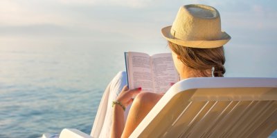 Pohled zezadu na ženu v klobouku a s knihou ležící na lehátku u moře.