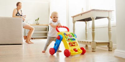 Dítě chodí s chodítkem, v pozadí sedí žena