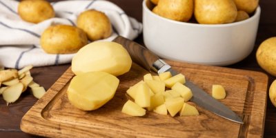 Z poloviny nakrájená brambora na dřevěném prkénku, položený nůž a v pozadí hrnec s bramborami