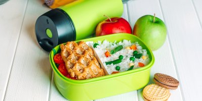 Otevřená svačinová krabička, ve které je rýže se zeleninou a pečivo, vedle jsou jablka, sušenky a termoska