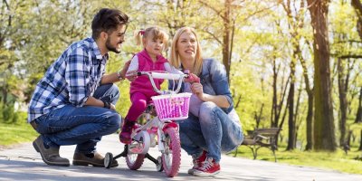 Holčička v parku sedí na dětském kole s balančními kolečky vedle svých rodičů 