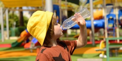 Chlapec v tričku a žlutém klobouku pije vodu z pet lahve na dětském hřišti 