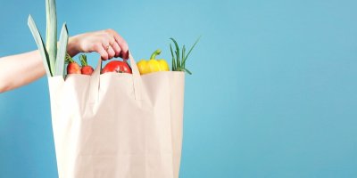 Ženská ruka drží papírovou tašku s potravinami, modré pozadí