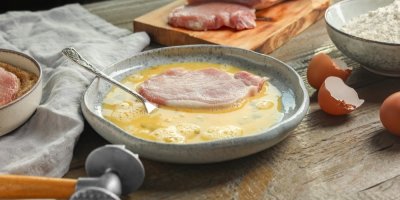 Syrový plátek masa v rozkvedlaném vajíčku, v pozadí maso, kladívko, skořápky