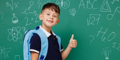 Chlapec se školním batohem na zádech stojí před školní tabulí a ukazuje palec nahoru