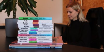 Učebnice fyziky, matematiky a literatury, v pozadí studentka sedící na kancelářské židli za stolem