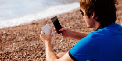 Muž sedí na pláži a nabíjí telefon přes powerbanku