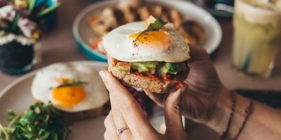 Žena drží v rukou toast s vejcem, v pozadí snídaňové menu