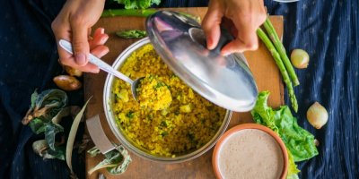 Pokrm z bulguru v hrnci, ženské ruce držící lžíci a pokličku, prkénko se zeleninou