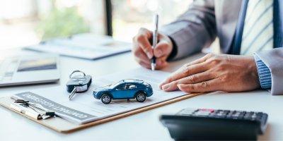 Ruce podepisují papír, na kterém leží klíče od auta a malé autíčko