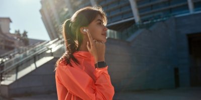 Mladá žena v oranžové větrové bundě, s chytrými hodinkami a s bezdrátovými sluchátky v uších