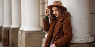 Žena v hnědém kabátě a klobouku se opírá o sloup.