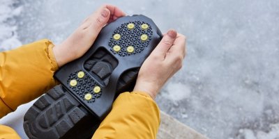 Člověk ve žluté bundě drží v rukou botu s protiskluzovým návlekem