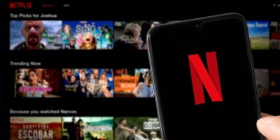 Logo Netflixu na obrazovce mobilního telefonu, v pozadí katalog filmů na platformě Netflix