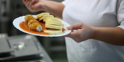 Kuchařka ve školní jídelně drží talíř s rajskou omáčkou, knedlíkem a masovými kuličkami