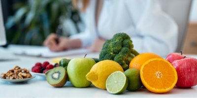 V popředí ovoce a zelenina, v pozadí žena něco píše