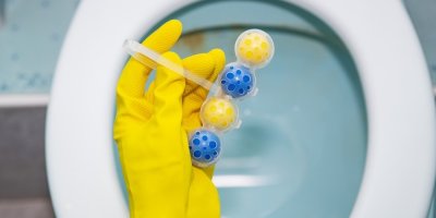 Ruka ve žluté rukavici drží závěsné WC kuličky nad záchodovou mísou