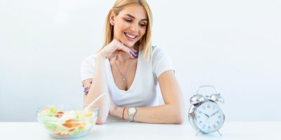 Žena sedí u stolu s miskou salátu a dívá se na budík